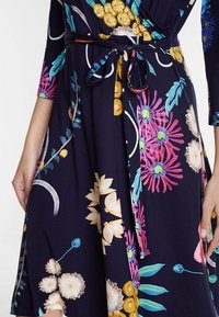 Květinové zavinovací šaty s tmavě námořnickým základem, zdobené barevnými květinami v růžové, žluté a modré, se šňůrkou v pase odpovídající barvy.