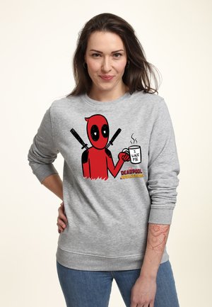 Vrouw met lang bruin haar draagt een grijze trui met Deadpool die een mok vasthoudt met de tekst "I like me", gecombineerd met een blauwe spijkerbroek.