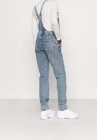 Vaaleansiniset farkkutakit, joissa on säädettävät olkaimet, sivutaskut ja takataskut. Yhdistetty valkoisiin urheilujalkineisiin, mikä korostaa rentoa istuvuutta.