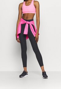 Mustat legginsit, vaaleanpunainen urheiluliivi, jossa on pieni logo, ja sidottu vaaleanpunainen paita. Kengät ovat tummat valkoisilla yksityiskohdilla. Materiaalit ovat sileät ja istuvat.