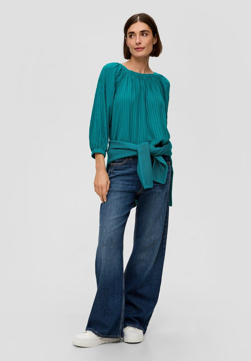 Türkisfarbige Bluse mit gefältelter Textur und gerafftem Kragen, kombiniert mit dunkelblauen, weit geschnittenen Jeans und weißen Sneakers.