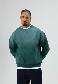 Groene trui met een losse pasvorm, voorzien van decoratieve zwarte stippen en een ronde hals. Gecombineerd met donkerblauwe spijkerbroek.