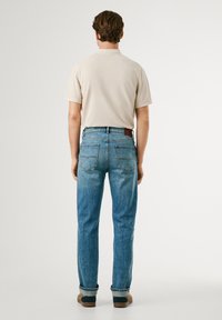 Camisa polo de color beige claro, jeans de denim azul con un ligero desvanecimiento, corte recto y parche de cuero marrón en la cinturilla, combinados con zapatos marrones.