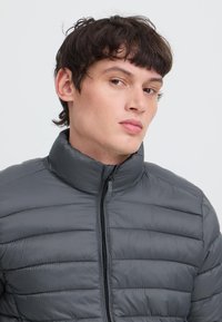 Mladý muž s krátkymi tmavými vlasmi, oblečený v sivej prešívanej bunde na zips s stojatým golierom, ktorý sa jemne pozerá do strany na jednofarebnom pozadí.
