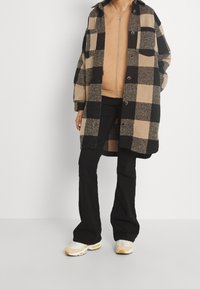 Una persona está de pie vistiendo un gran abrigo a cuadros negro y marrón claro sobre una camiseta de cremallera marrón claro, combinado con pantalones negros y zapatillas beige.