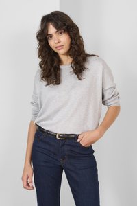 Svetlo siva oversized pulover z rebrastimi manšetami, v kombinaciji s temno modrimi jeans hlačami z oranžnim šivom in črnim pasom z zlatim sponko.