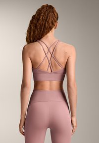 Femeie cu păr lung și creț, purtând un top sportiv mov cu bretele încrucișate și colanți cu talie înaltă, stând cu spatele spre cameră.