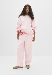 Felpa oversized rosa chiaro con un accento a righe rosse sull'orlo, abbinata a pantaloni larghi rosa coordinati e sneakers beige.