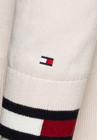 Strickpullover in Creme mit gerippten Bündchen, der ein horizontales Streifenmuster in Rot, Weiß und Marineblau aufweist. Kleines Logopatch am Ärmel.