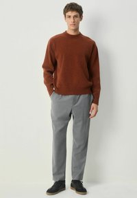 Ein junger Mann steht gegen eine weiße Wand und trägt einen rostfarbenen Pullover, graue Hosen und schwarze Sneaker mit Gummisohlen.