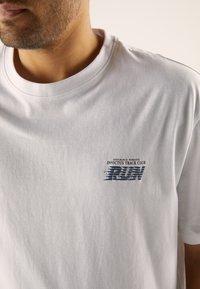 White cotton t-shirt featuring a round neckline, short sleeves, and a printed graphic in blue and black on the left chest.