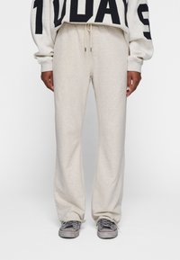Pantalones de chándal beige de corte holgado, hechos de material suave. Presentan una cintura con cordón y sutiles detalles en las costuras laterales. Combinados con zapatillas grises.