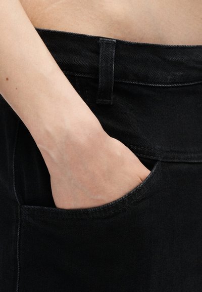 Χέρι τοποθετημένο μέσα στην μπροστινή τσέπη μαύρου τζιν ψηλόμεσου με ορατή θηλιά ζώνης και ραφή.