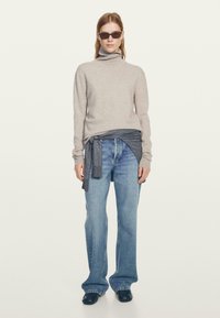 Maglione a collo alto grigio chiaro, strato grigio scuro legato in vita, jeans a gamba svasata blu, calzature nere e occhiali da sole scuri. Sfondo semplice.