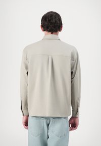 Camicia beige chiaro a maniche lunghe con colletto, piega centrale sul retro e polsini abbottonati, abbinata a jeans in denim blu chiaro.