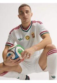 Jeune joueur de football masculin en tenue blanche avec des bordures rouges et vertes, accroupi tenant un ballon de football coloré sur fond blanc.
