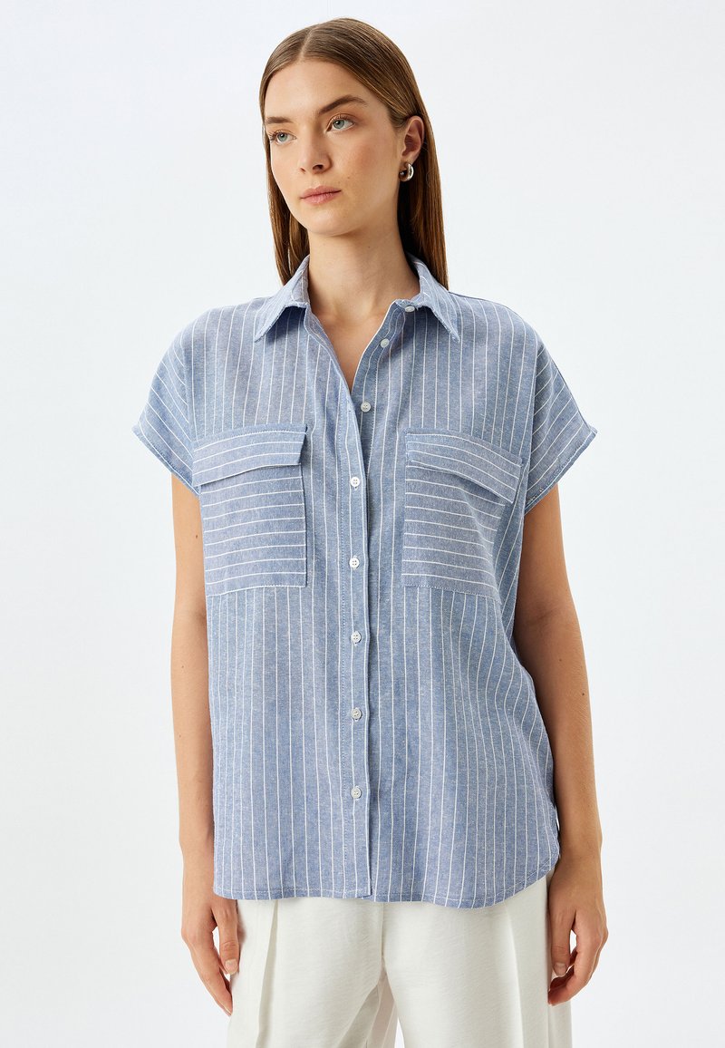Camisa de manga corta azul claro con rayas verticales blancas, dos bolsillos en el pecho y cierre de botones en la parte delantera. Fabricada con un tejido suave y de corte relajado.