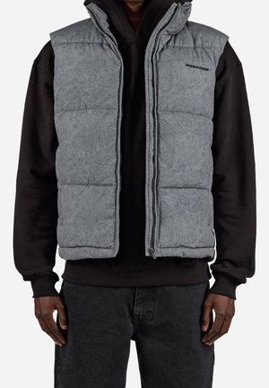 Homme portant une veste matelassée sans manches grise par-dessus un sweat à capuche noir et un jean foncé, debout avec les bras détendus le long du corps.