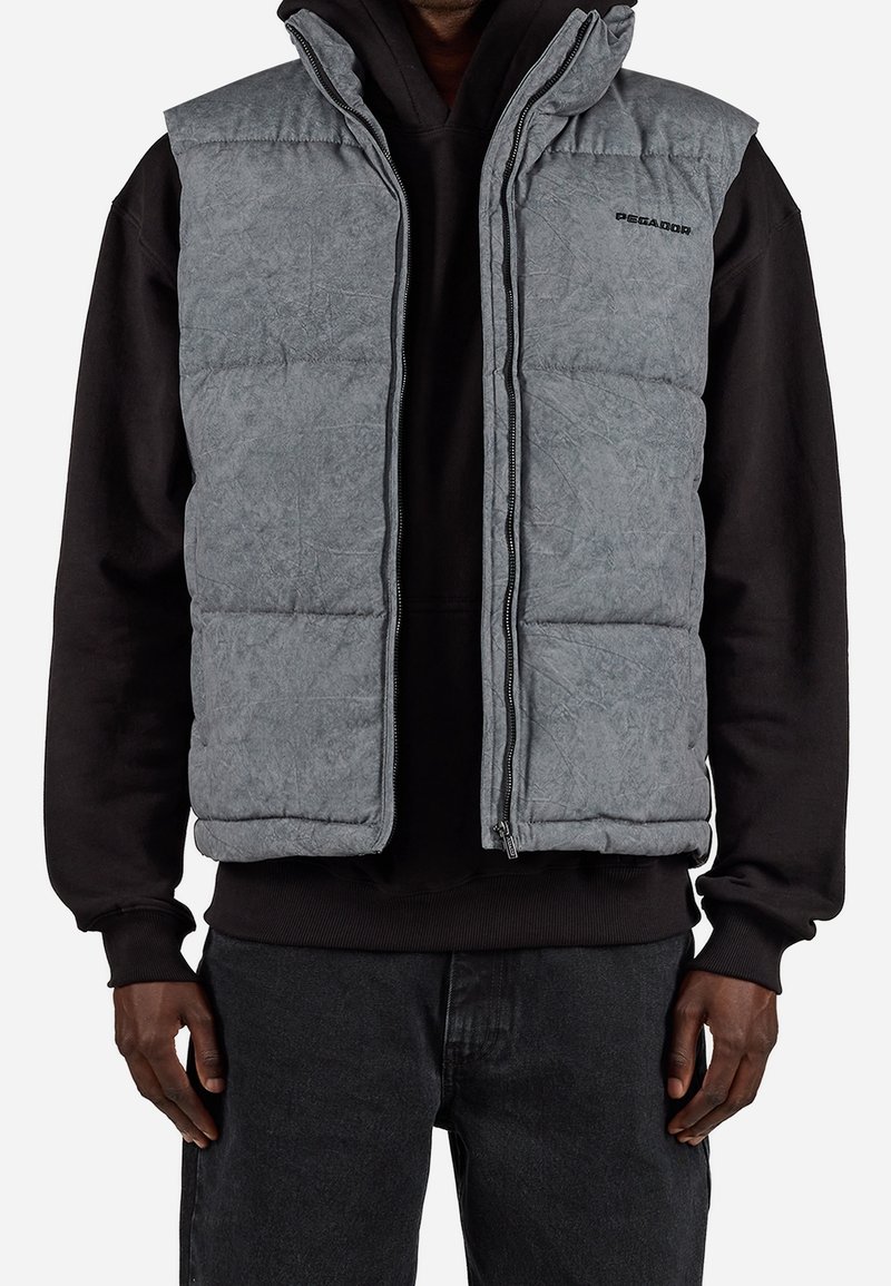 Homme portant une veste matelassée sans manches grise par-dessus un sweat à capuche noir et un jean foncé, debout avec les bras détendus le long du corps.