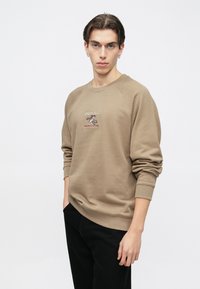 Καφέ φούτερ με ραγκλάν μανίκια, κεντημένο λογότυπο στο στήθος, ριμπ μανσέτες και τελείωμα· φορεμένο με μαύρο παντελόνι.