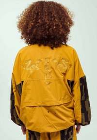 Chaqueta amarilla con dragones bordados y logotipo, con un corte holgado, mangas negras con estampado y un tejido ligero y texturizado.