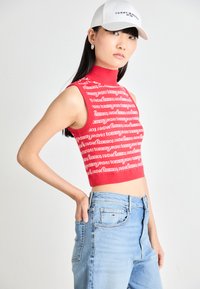 Raudona liemenė be rankovių, padengta pakartotine balta "TOMMY JEANS" logotipu, derinama su šviesiai mėlynomis džinsais. Modelis taip pat dėvi baltą kepurę ir sidabro auskarus.