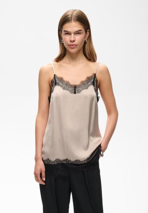Tânără cu părul drept, purtând o camisola din satin bej cu margini din dantelă neagră și pantaloni negri, stând cu o mână în buzunar.