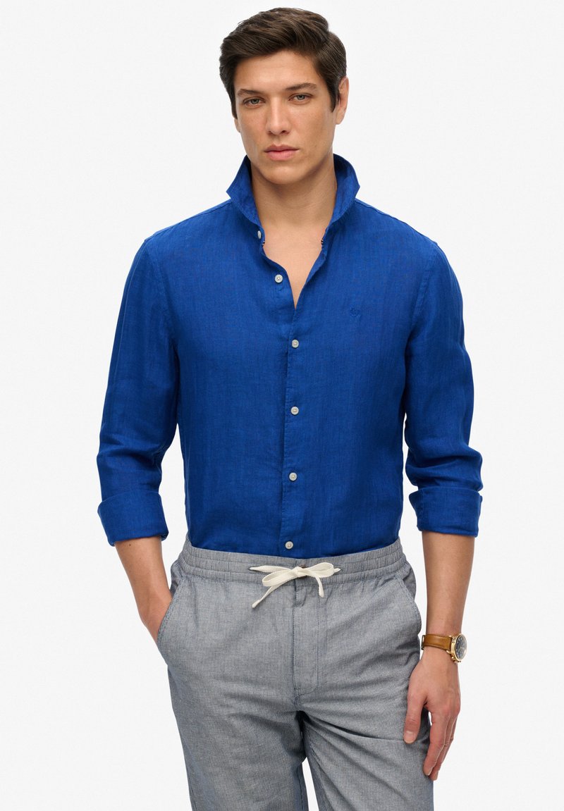 Camisa de lino azul con cuello abotonado, mangas enrolladas y un logo sutil. Combinada con pantalones elásticos de cintura gris con rayas y un reloj.
