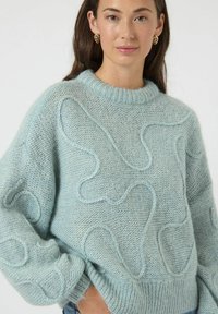 Jasnoniebieski sweter z dzianiny o miękkiej teksturze, z podniesionymi falistymi wzorami i ściągaczem przy dekolcie. Luźny krój z szerokimi rękawami.
