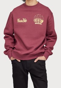 Sudadera burdeos con puños y dobladillo acanalados, que presenta diseños gráficos dorados en el pecho; textura suave, corte relajado.