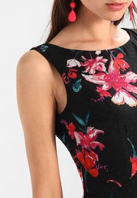 Musta hihaton mekko, jossa on kirkkaan punaisia ja teal-värisiä kukkakuoseja. Teksturoitu kangas, jossa on koholla olevia pisteitä ja pyöreä kaula-aukko.