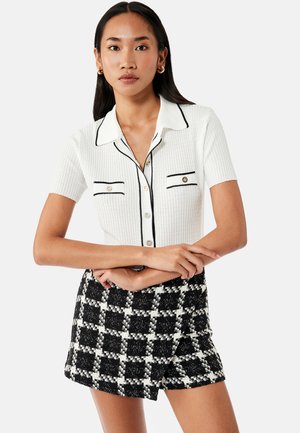 Mujer con camiseta de punto blanca de manga corta con ribete y botones negros, combinada con pantalones cortos a cuadros en blanco y negro, de pie con los brazos cruzados.