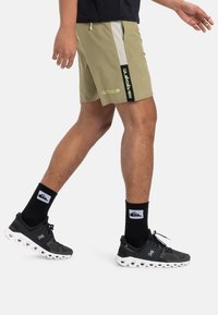 Pantalones cortos deportivos caqui con acentos en negro y gris, que cuentan con un logo. Combinados con zapatillas negras y calcetas altas negras.