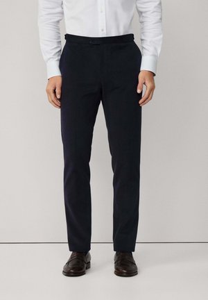 Pantalon marine avec une texture lisse, coupe slim, fermeture à bouton et passants de ceinture, associé à des chaussures de ville foncées.
