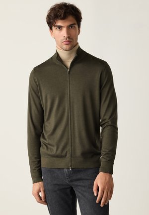 Mann trägt einen dunkelgrünen Reißverschluss-Pullover über einem beigen Rollkragenpullover mit dunklen Jeans vor einem schlichten hellen Hintergrund.