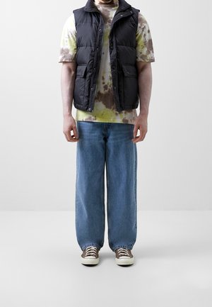 Zwart gewatteerd vest over een tie-dye T-shirt met korte mouwen in geel, bruin en grijs, gecombineerd met losse blauwe spijkerbroeken en bruine sneakers.