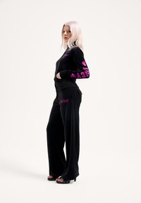 Fekete velúr melegítő hosszú ujjakkal, rózsaszín mintákkal az ujjakon és a csípőn. Laza fazonú nadrágok, enyhén bővülőek, sima tapintásúak.