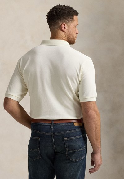 Šviesiai smėlio spalvos polo marškinėliai su trumpomis rankovėmis ir briaunotu apykakle, derinami su tamsiai mėlynais džinsais ir rudos odos diržu.