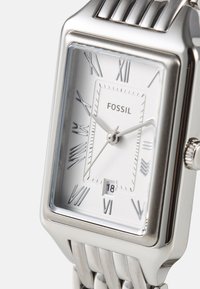 Montre rectangulaire argentée avec un boîtier en acier inoxydable poli, un cadran blanc, des index en chiffres romains, et un bracelet en métal avec un design strié.