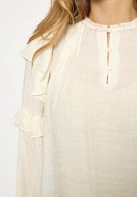 Kreemikas pluus, valmistatud kerge kangaga. Omaduseks volangidega varrukad, ümmargune kaelus ja ninakujuline avaus kolme nööbiga.