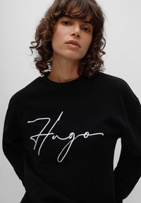 Schwarzes Sweatshirt aus Baumwolle mit Rundhalsausschnitt, das ein auffälliges weißes, geschwungenes "Hugo"-Logo über der Brust trägt.