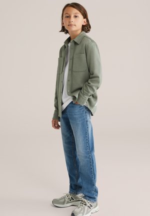 Grüne Hemdbluse mit zwei Brusttaschen, über einem weißen T-Shirt getragen, kombiniert mit hellblauen Jeans und grauen Sneakers mit Akzenten.