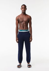 Navy sweatpants with elastic cuffs and a Lacoste waistband featuring green and white branding. Worn with black slide sandals.