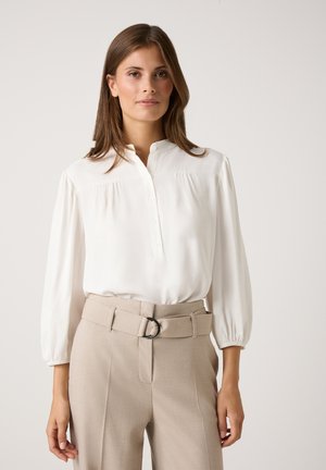 Weiße Bluse mit langen Ärmeln und Mandarin-Kragen, mit durchgehender Knopfleiste und Raffung. Kombiniert mit beigen, hoch taillierten Hosen.