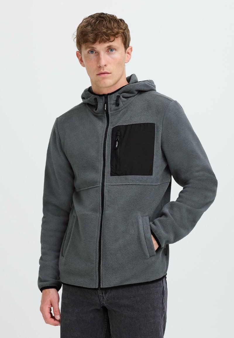 Junger Mann, der eine graue Fleecejacke mit Reißverschluss und einer schwarzen Brusttasche sowie Kapuze trägt, im Stehen vor einem schlichten hellen Hintergrund.