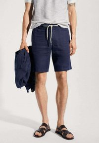 Shorts en lin bleu marine avec une taille élastique et un cordon de serrage. Associés à une chemise à manches courtes rayée et des sandales noires à enfiler.