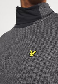 Pull-over gris foncé en tissu texturé avec un col montant, arborant un logo oiseau jaune sur le côté gauche. Comprend des accents noirs contrastants.