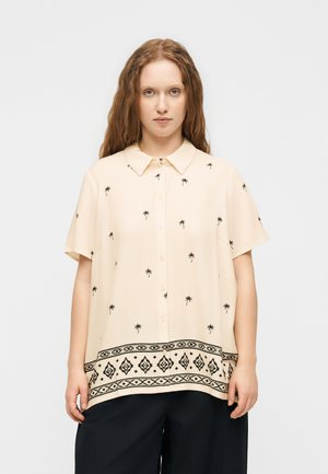 Joven con el pelo largo y ondulado que lleva una camisa cremosa de manga corta con pequeños estampados de palmeras y dobladillo negro estampado, y pantalones negros.