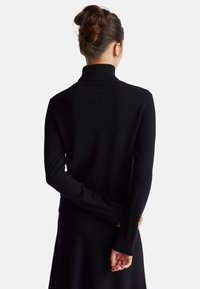 Frau mit braunen Haaren im Dutt, die einen schwarzen Rollkragenpullover und einen schwarzen Rock trägt, steht mit den Händen hinter dem Rücken und schaut weg.