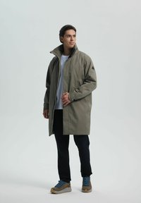 Oliwkowy wodoodporny płaszcz z wysokim kołnierzem, bocznymi kieszeniami i smukłym krojem, w połączeniu z ciemnymi spodniami i brązowymi butami trekkingowymi.